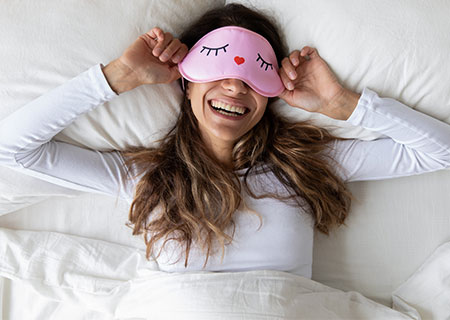 Lachende Frau im Bett mit Lustiger Schlafmaske - gut gelaunt im weichen Bett aufwachen